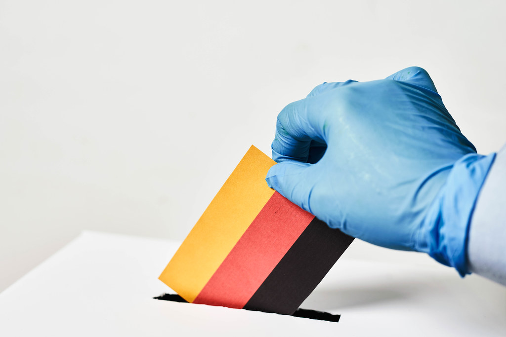 Für den Bundestag in Corona-Zeit abstimmen: Hand mit Einmalhandschuh wirft deutsche Flagge in Wahlurne ein