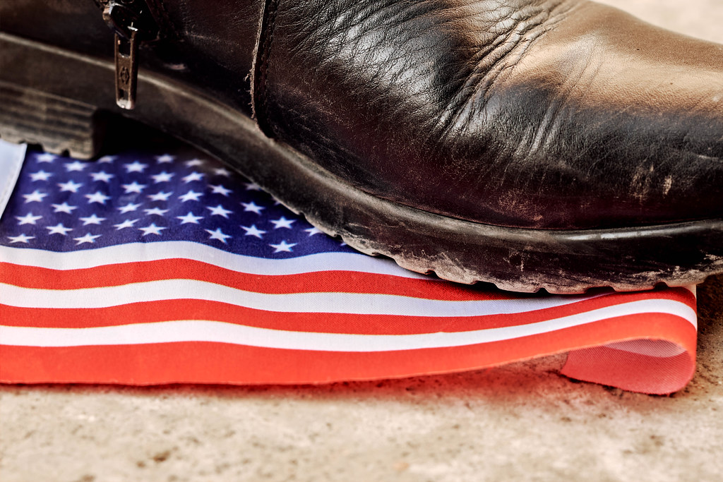 Fuß in einem schwarzen, dick besohlten Stiefel tritt mit Verachtung auf eine amerikanische Flagge