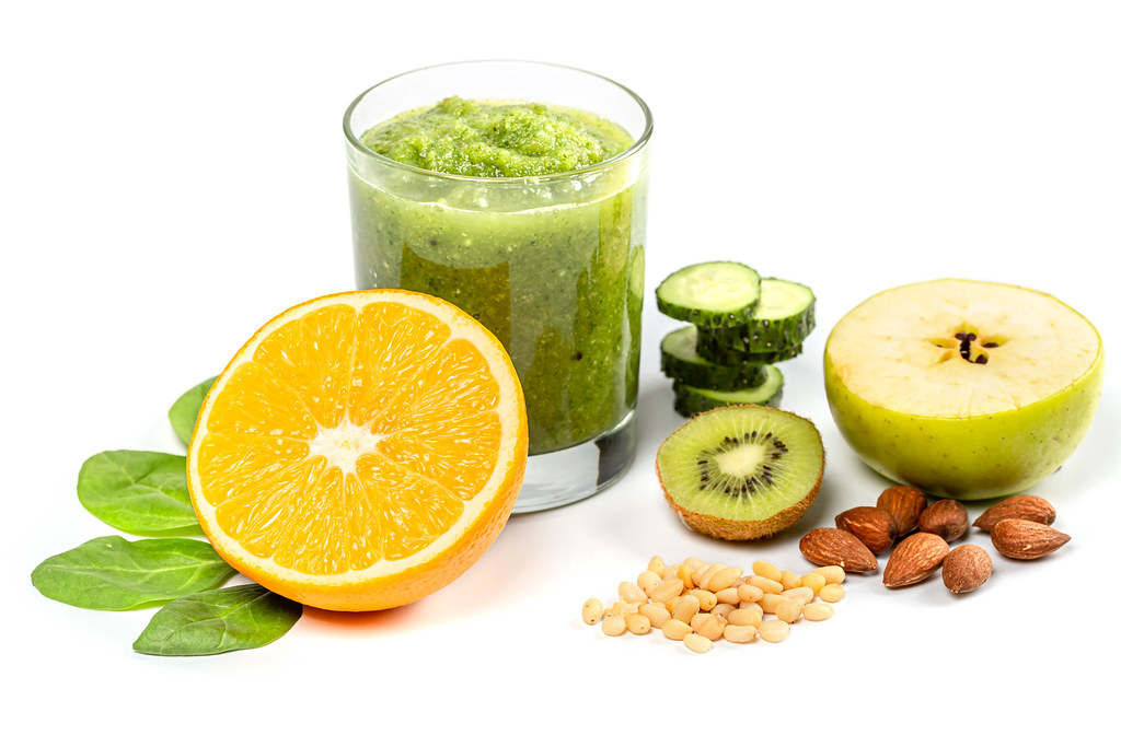 Glass of green smoothie with fresh apple, orange halves, sliced cucumber, spinach leaves, almonds and pine nuts