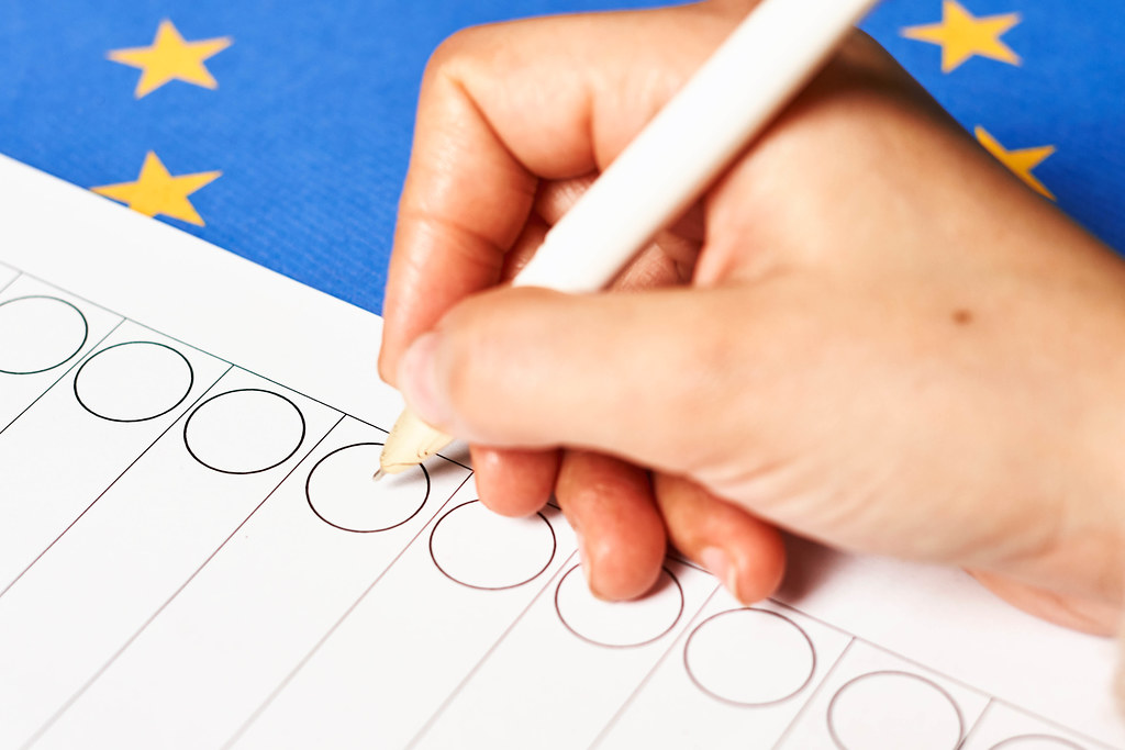 Hand eines Wählers mit dem Kugelschreiber macht ein Kreuz auf dem Stimmzettel vor der EU-Flagge