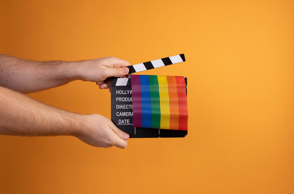 Hand holding movie clapper and rainbow flag on orange background