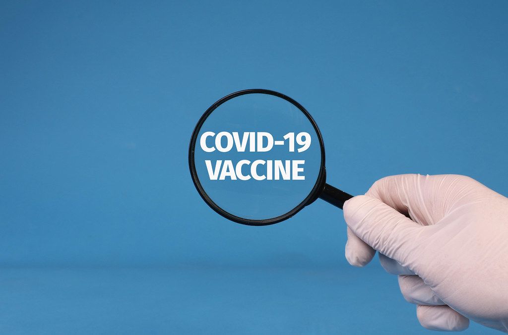 Hand in medical gloves holding magnifying glass over Covid-19 Vaccine text on blue background