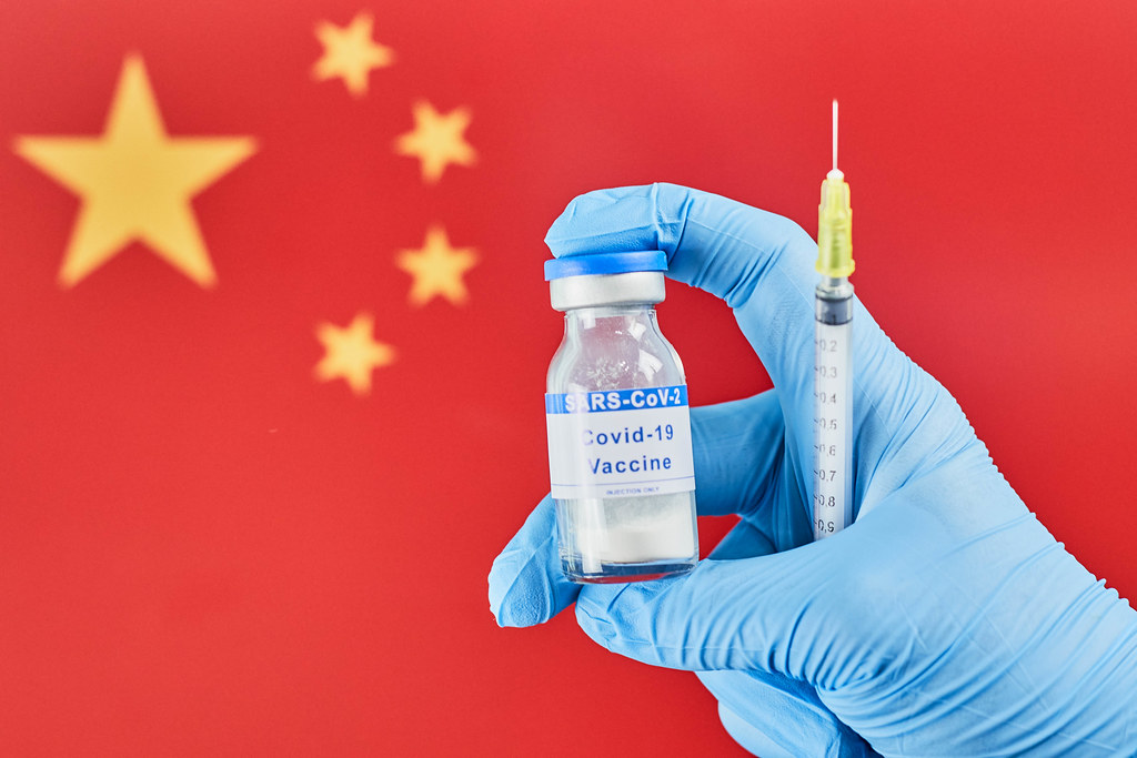 Hand in medical gloves holds Covid-19 vaccine and syringe against Chinese flag