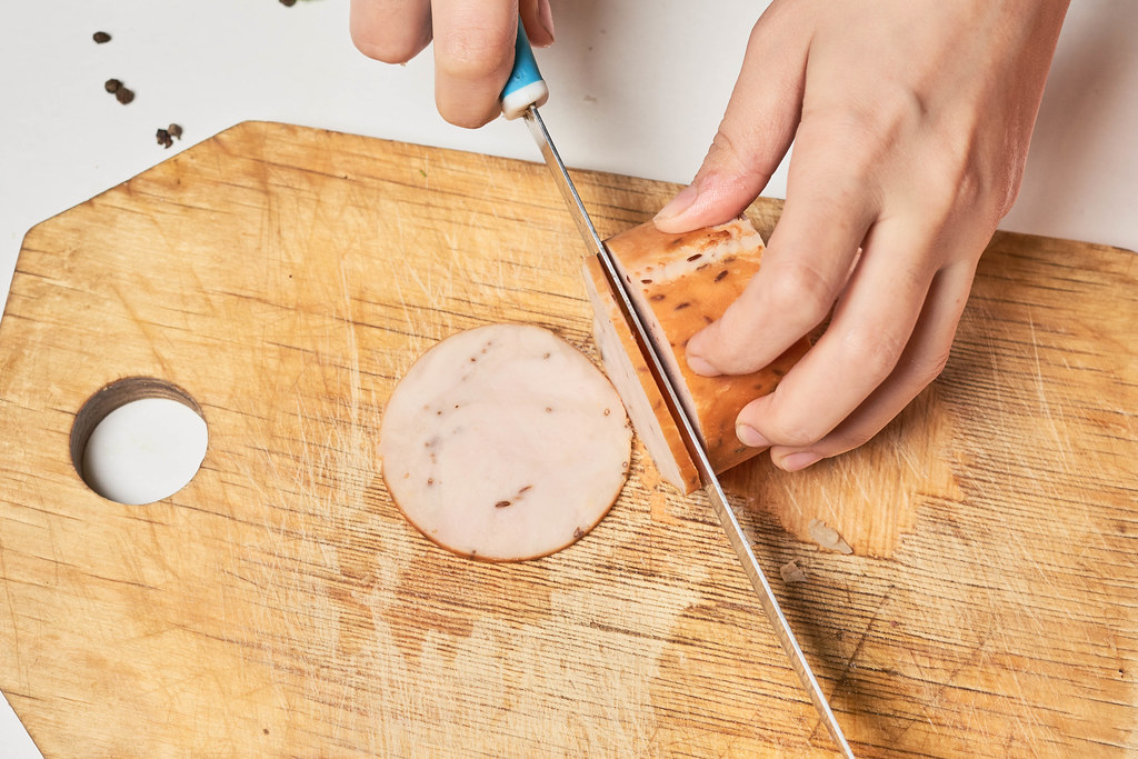 Hand schneidet eine geräucherte Hähnchen-Fleischwurst mit dem Messer auf einem Holzbrett
