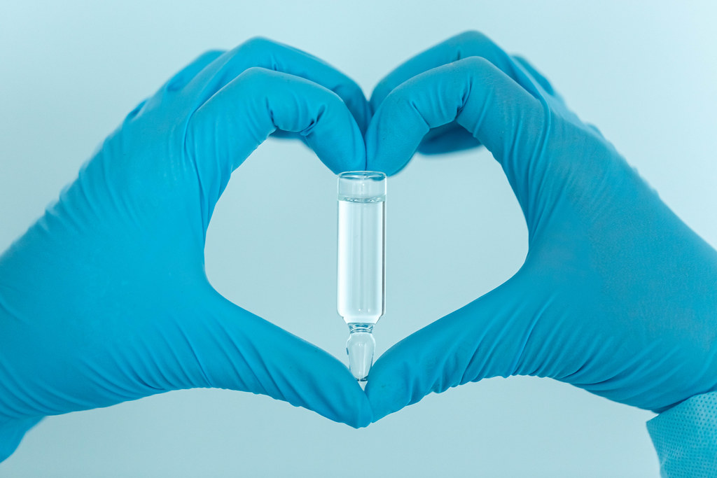 Hands in medical gloves form a heart shape with vaccine ampoule