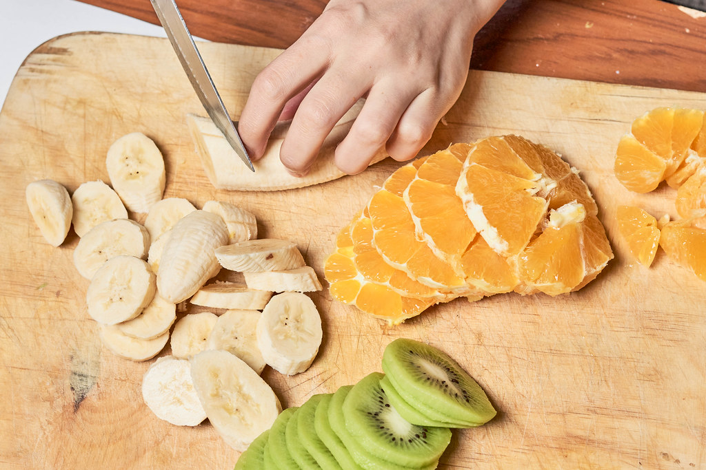 Holzbrett mit geschnittenen Kiwis, Orangen und Bananen. Hand mit Messer schneidet die Banane