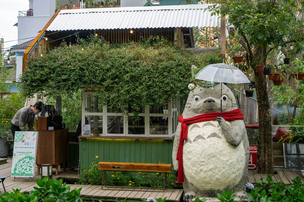 Human Size Japanese Character Totoro with Red Scarf and Transparent Umbrella in front of a Wooden House at Still Cafe in Da Lat, Vietnam