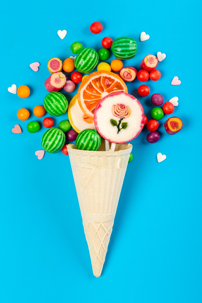 Ice cream waffle cone with colorful lollipop on stick, scattering of multicolored sweets and confectionery topping