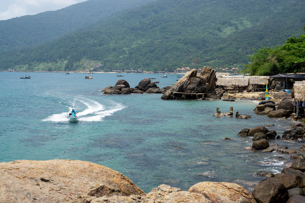 Jet Ski driving towards large Rocks at a Beach with Restaurant and Beach Bar with National Park in the Background on Son Tra Peninsula in Da Nang, Vietnam