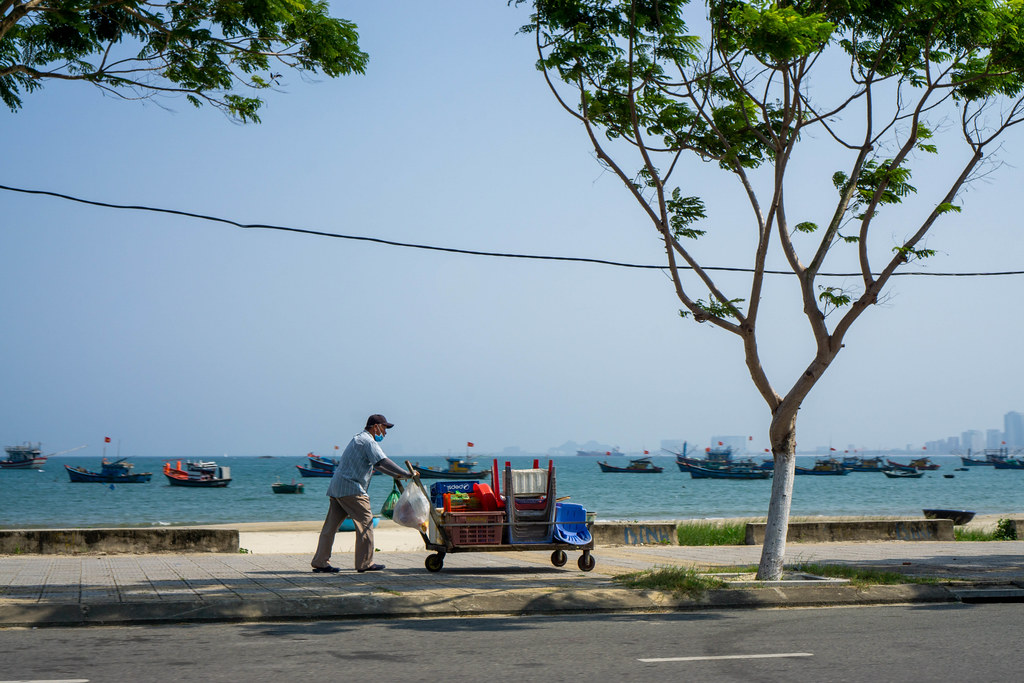 Man pushing a Street Food Cart with Plastic Chairs and Tables on a Sidewalk with Fishing Boats in the East Vietnam Sea in the Background in Da Nang, Vietnam