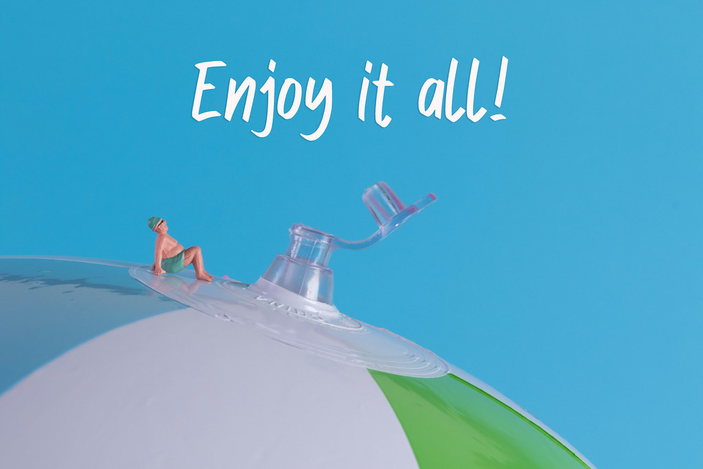 Miniature man in swimsuit sitting on a beach ball with Enjoy it all text