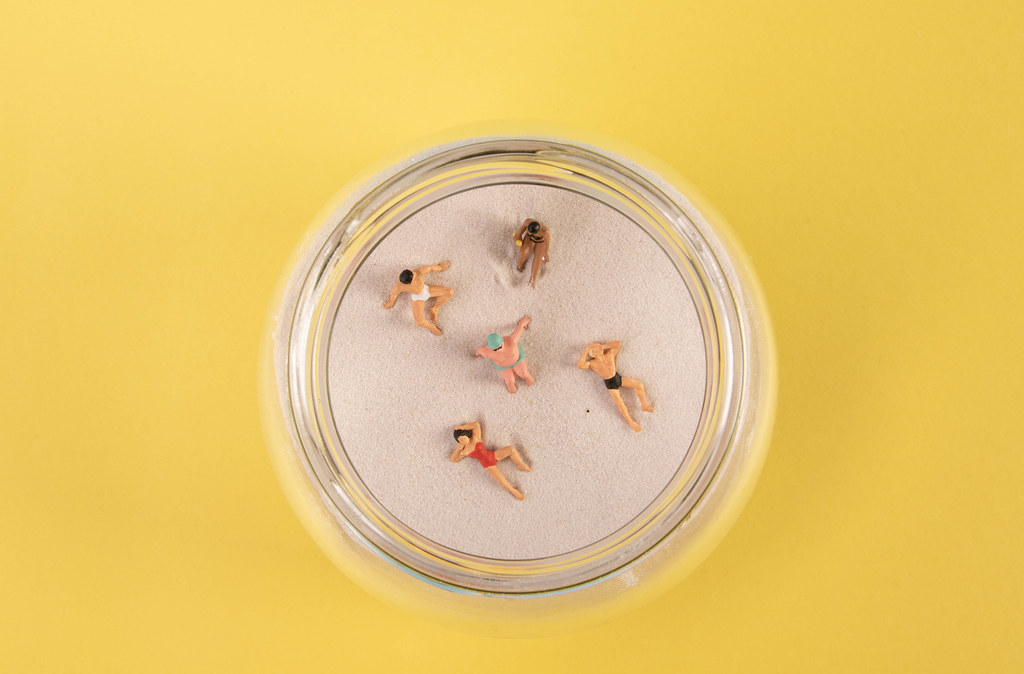 Miniature people relaxing on the sand in glass jar on yellow backgorund