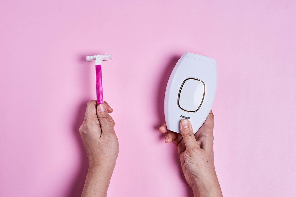 Modern laser depilator and razor in woman hands on over pink background