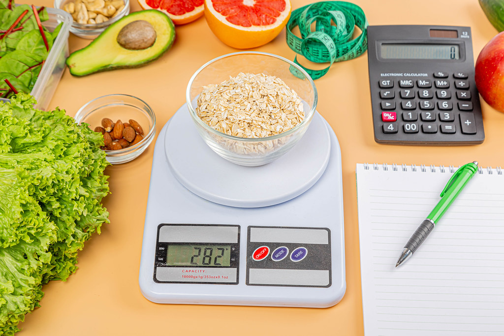 Oat flakes on kitchen scale on orange background with calculator, measuring tape and healthy foods