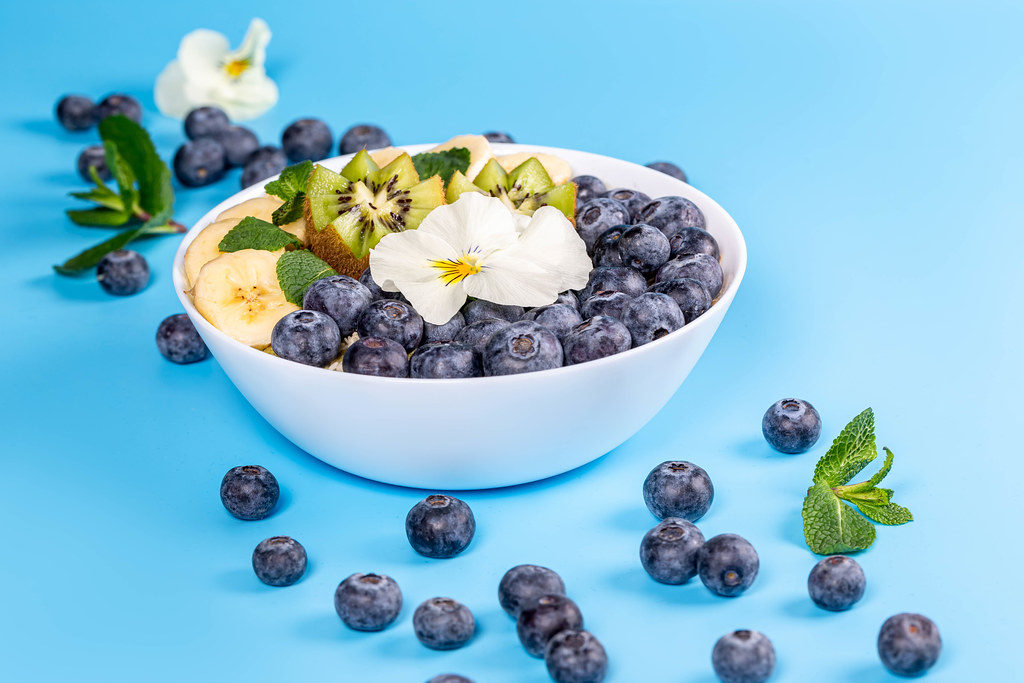 Oatmeal with blueberry, banana, kiwi and flower on blue background with fresh blueberries and mint leaves