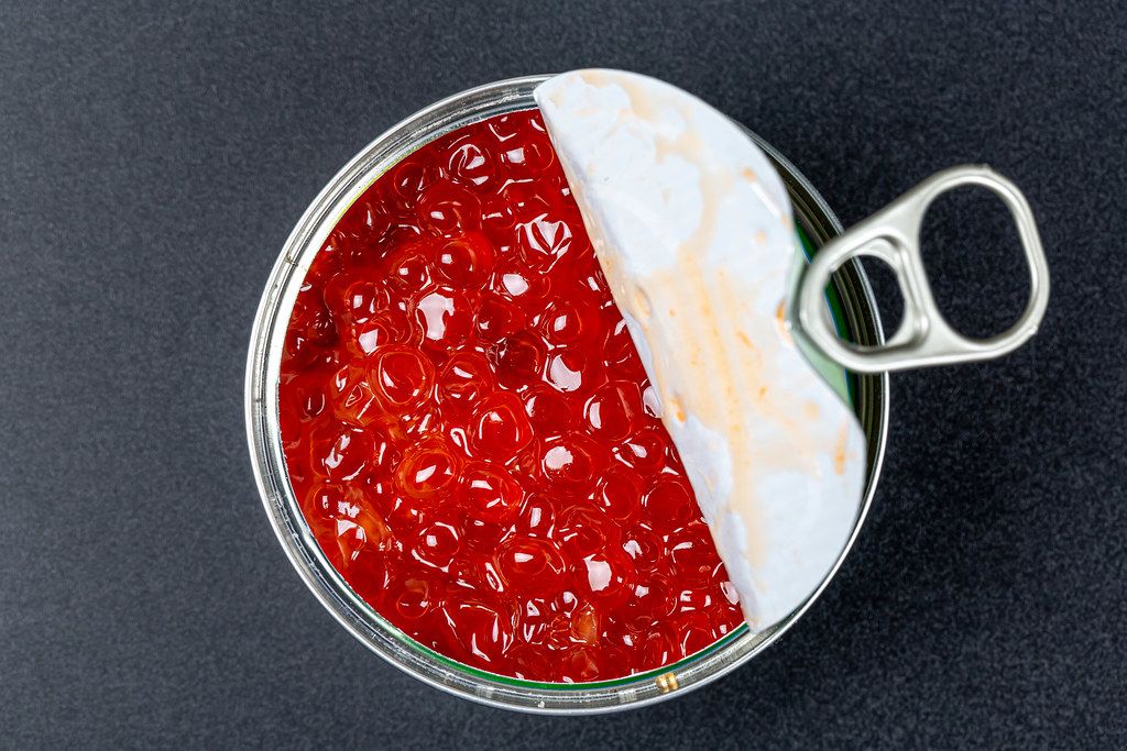 Open iron jar with red caviar on a black background, top view
