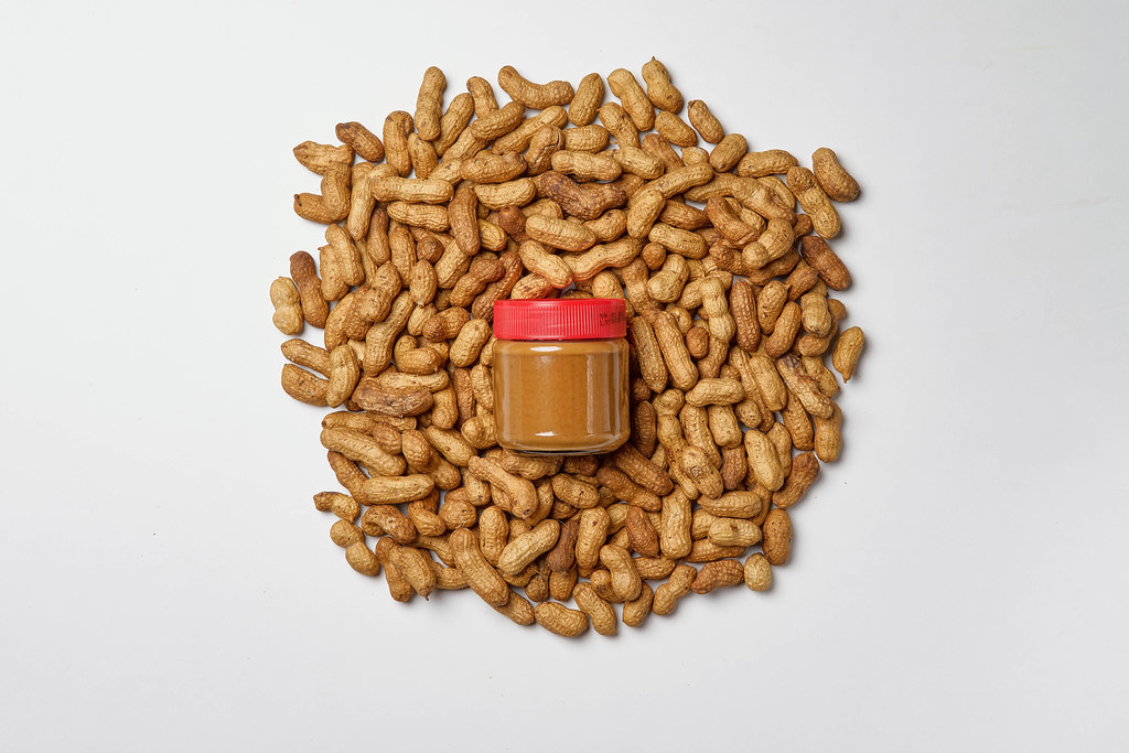 Peanut cream in glass jar with pile of whole nuts on white background