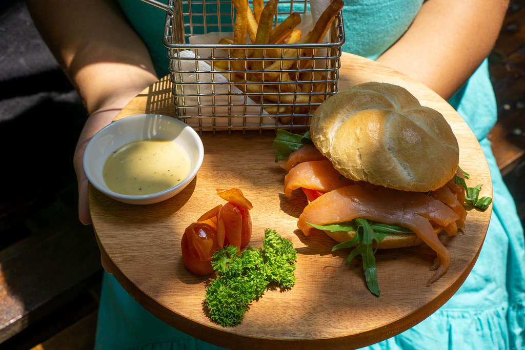Person holding a Wooden Board with Smoked Salmon Sandwich with Arugula in a German Bread Roll, Honey Mustard Sauce, Tomatoes, Parsley and Homemade French Fries in a Basket