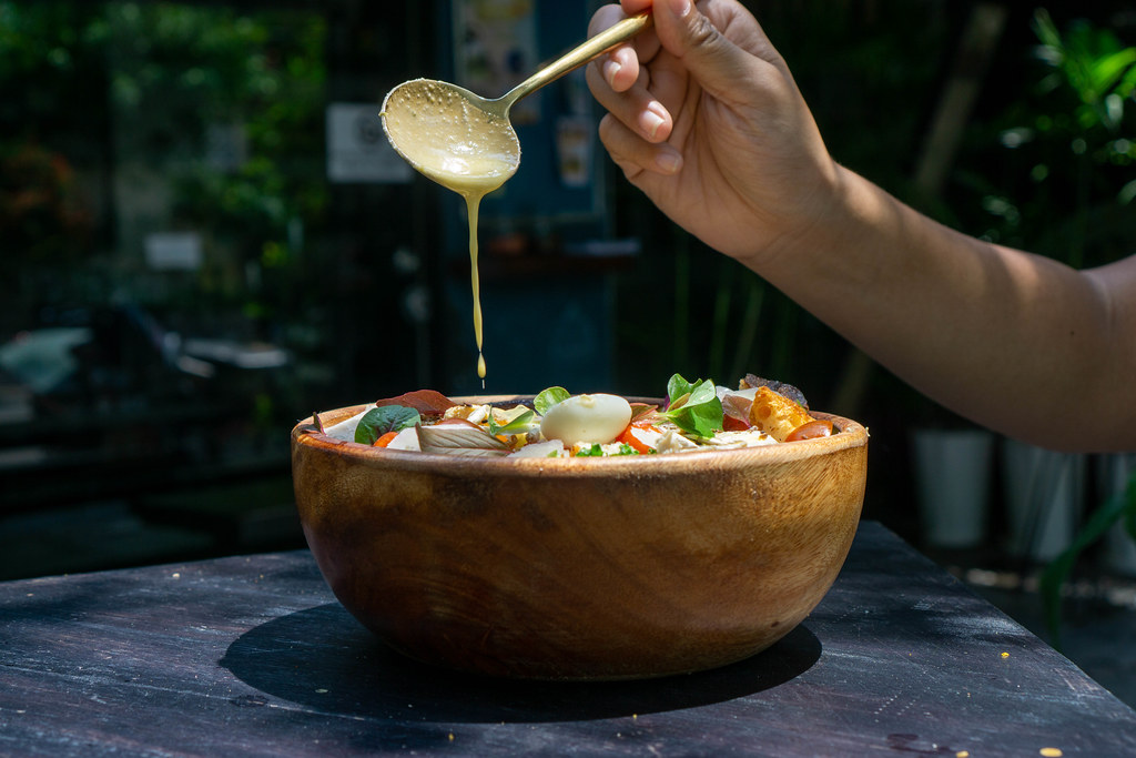 Person is pouring Honey Mustard Sauce over a Mixed Salad Bowl with Basil, Boiled Quail Eggs, Chicken and Parmesan Cheese with a Spoon in an Outdoor Space of a Restaurant