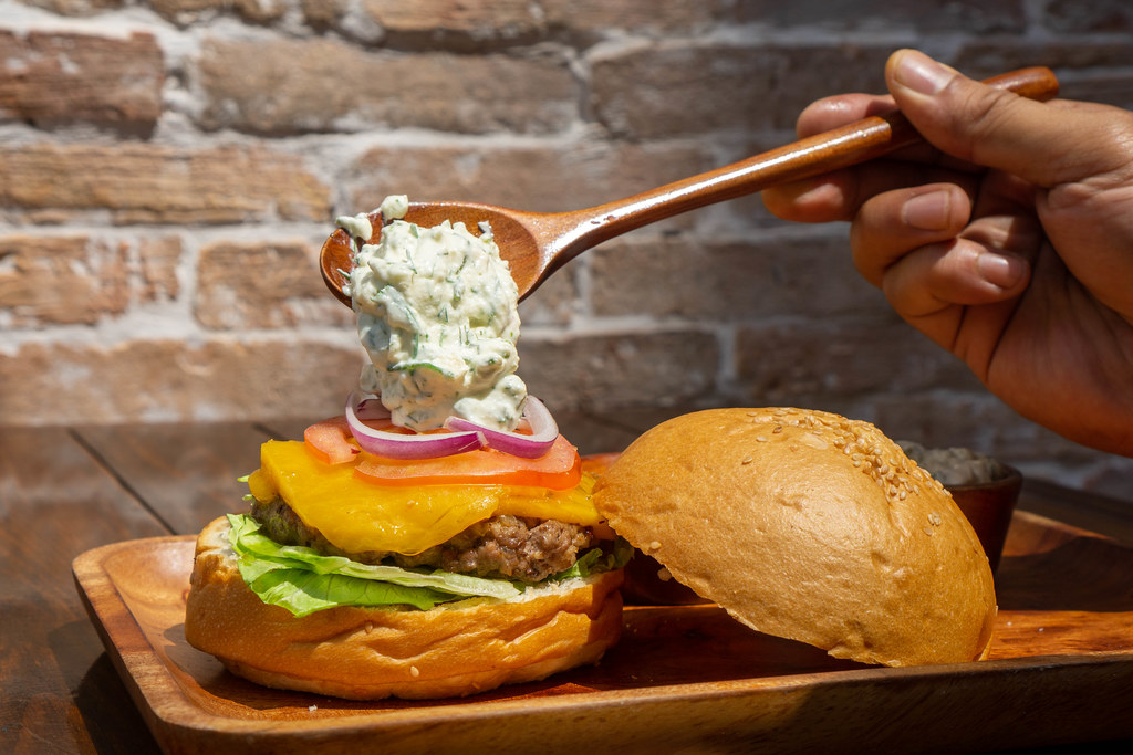 Person using a Wooden Spoon to put Tzatziki Sauce on a Lamb Kofta Burger with Lettuce, Cheese, Onions and Tomato in an Outdoor Restaurant