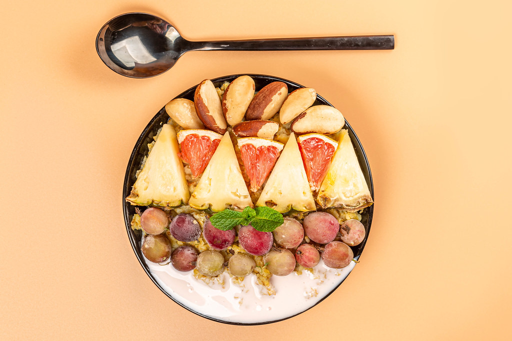 Porridge with brazil nuts, pineapple slices, grapefruit, gooseberries and yogurt on an orange background, top view