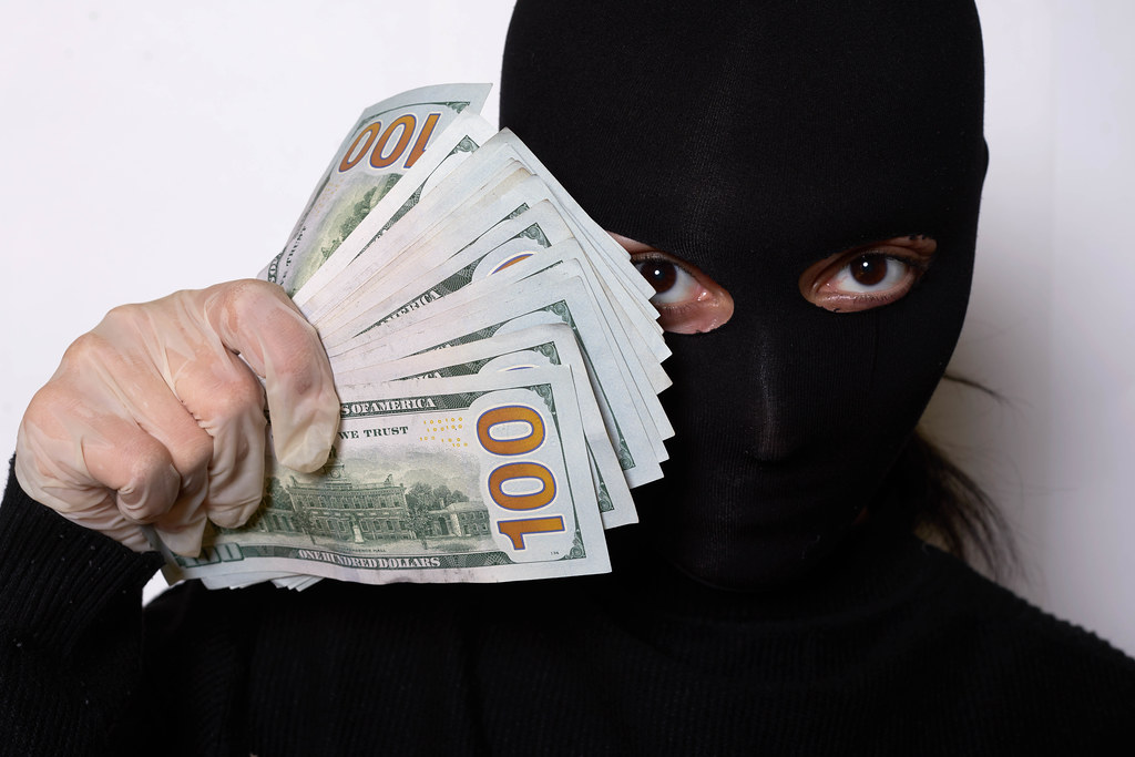 Portrait shot of a thief woman in black face mask holding a pile of stolen US dollars