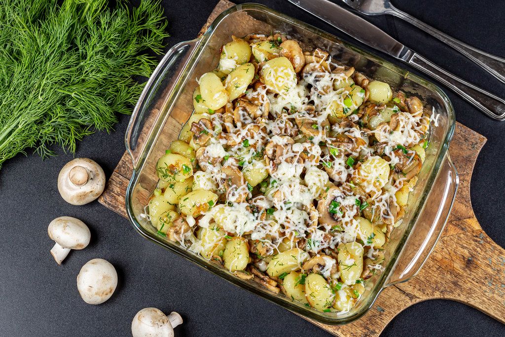 Potato gnocchi in a glass pan with mushrooms and cheese on a dark background, top view