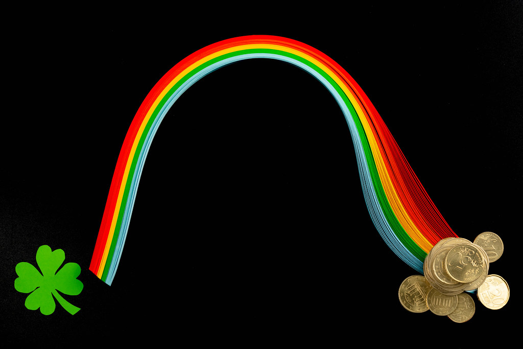 Rainbow from multi-colored paper with coins and paper clover leaf on a black background