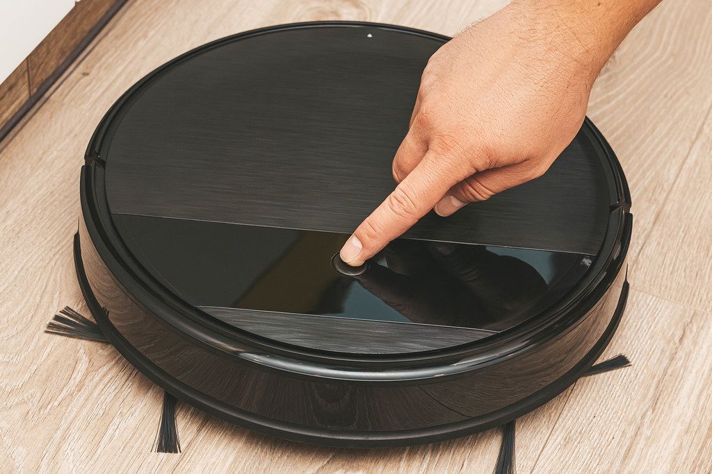 Robotic vacuum cleaner on laminate floor in living room, man presses button to turn on