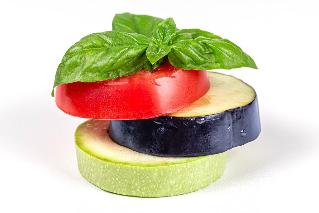 Sliced fresh vegetables - eggplant, zucchini, tomato with basil leaves on a white background