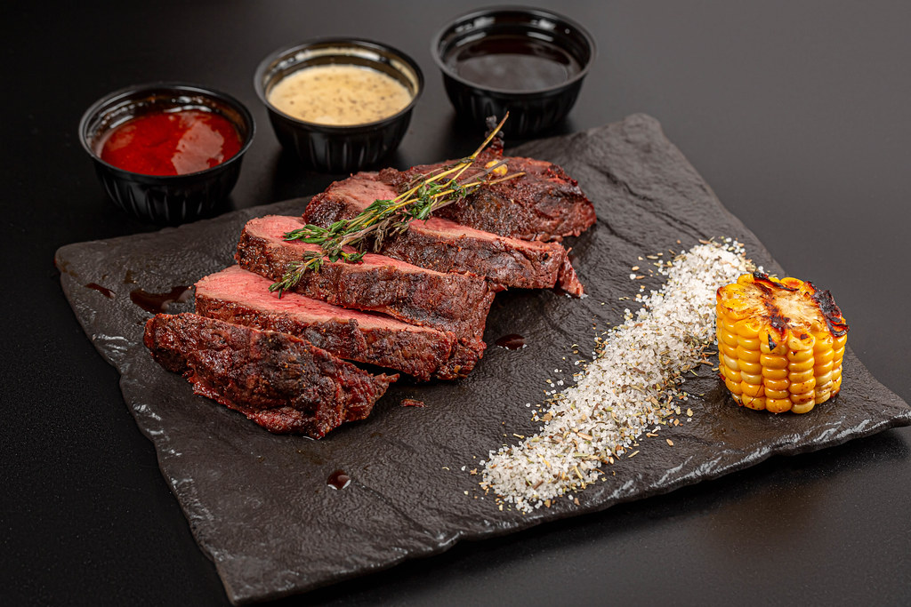 Sliced pieces of grilled beef with spices, thyme and sauces on a dark background