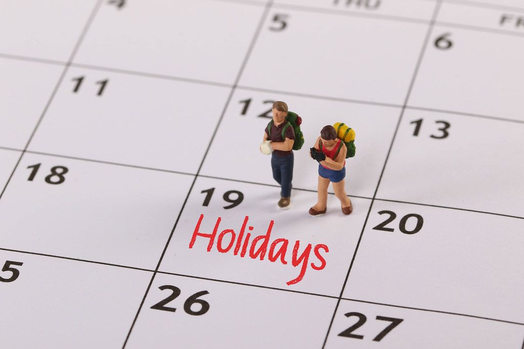 Small traveler figures with backpack standing on calendar with Holidays text
