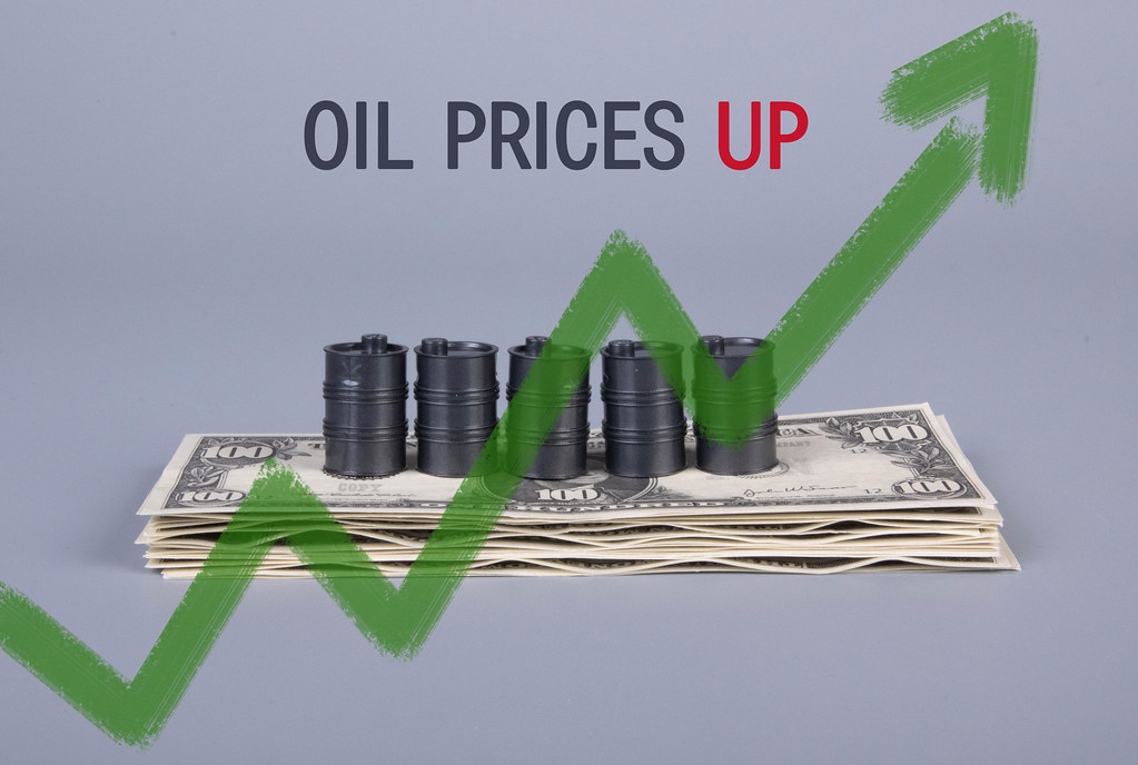 Stack of money and oil barrels with green up chart and Oil Prices Up text