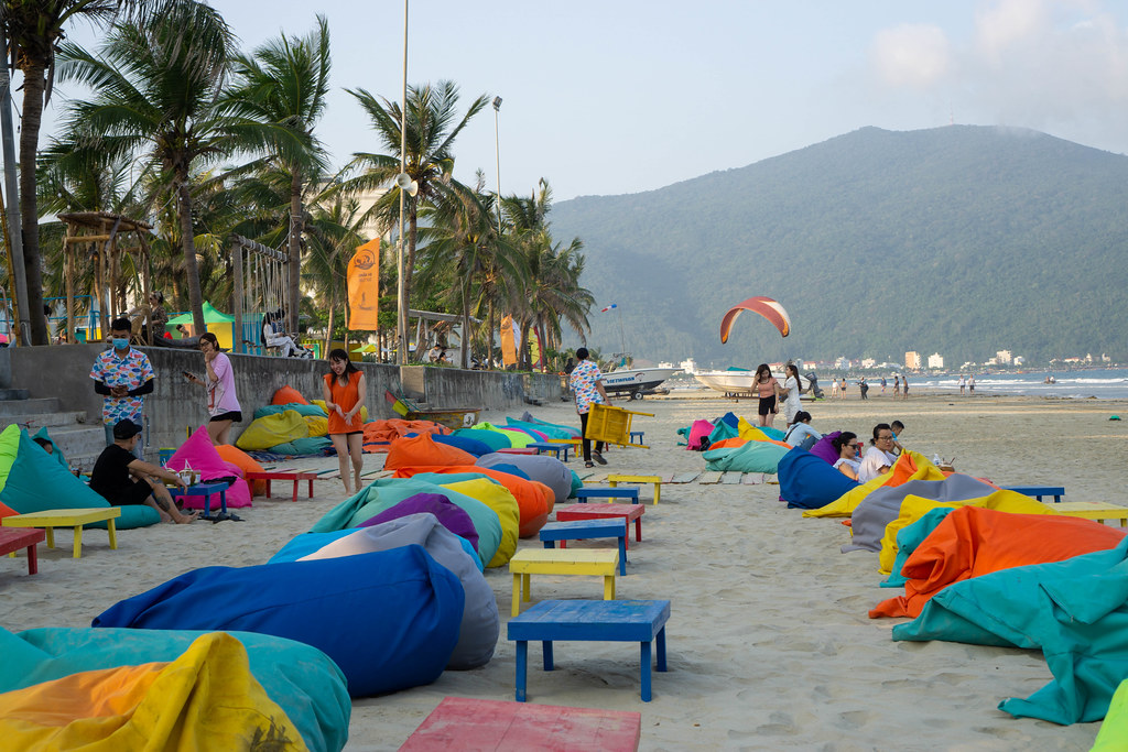 Strandbar mit Sandsäcken und Holztischen in vielen Farben mit Son Tra Halbinsel und Palmen im Hintergrund in Da Nang, Vietnam