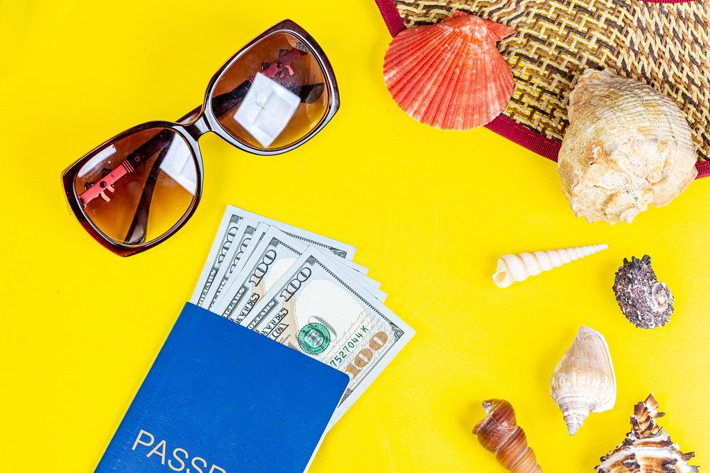 Straw hat, sunglasses, shells with passport and money on yellow background