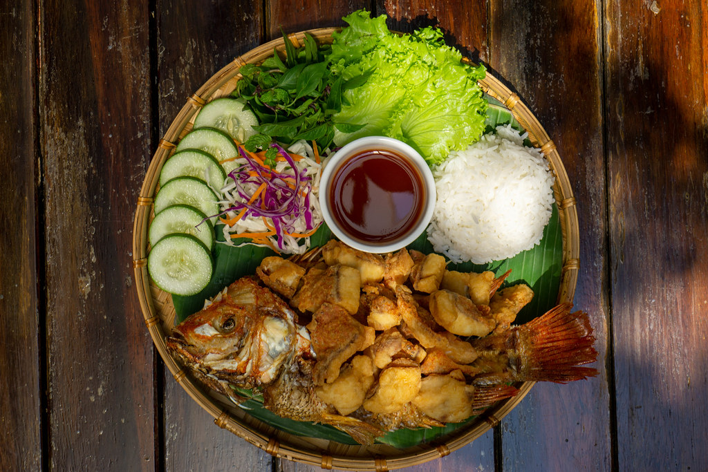 Top View Food Photo of Fried Tilapia Fish on a Bamboo Plate with Sliced Cucumber, Mixed Salad, Rice and Tamarind Sauce at a Vietnamese Restaurant