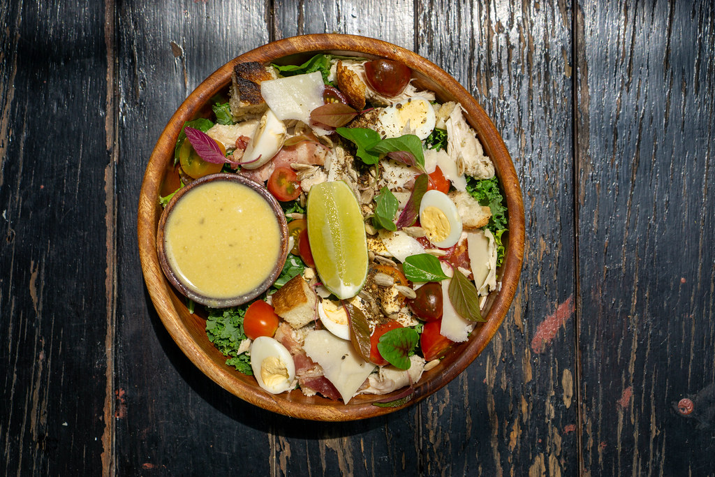 Top View Food Photo of Healthy Mixed Bowl with Boiled Chicken, Quail Eggs, Cherry Tomatoes, Parmesan Cheese, Basil, Lettuce, Lime and Honey Mustard Sauce on a Wooden Table