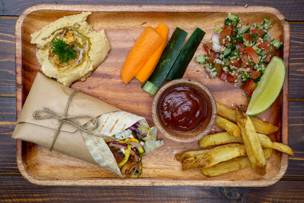 Top View Food Photo of Lunch Plate with Doner Durum Kebab Roll, Homemade Hummus Dip, Fresh Vegetables, Quinoa Salad and French Fries on a Wooden Board with Ketchup and Lime