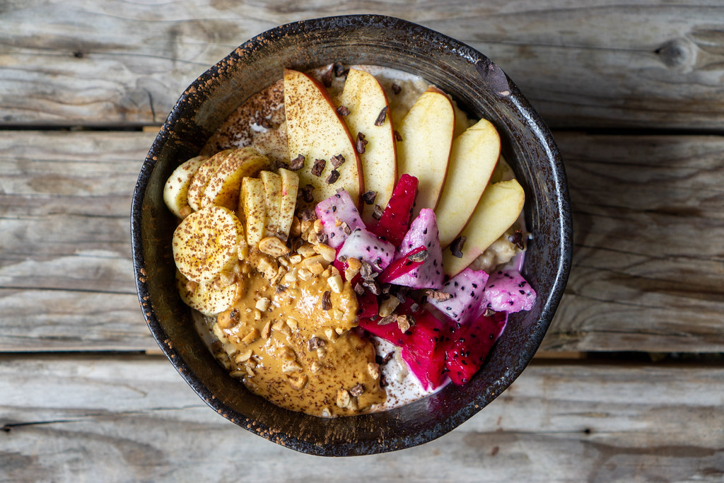 Top View Food Photo of Oatmeal Breakfast Bowl with Sliced Apple, Banana, Dragon Fruit, Coconut Milk, Peanuts and Peanut Butter on a Wooden Table