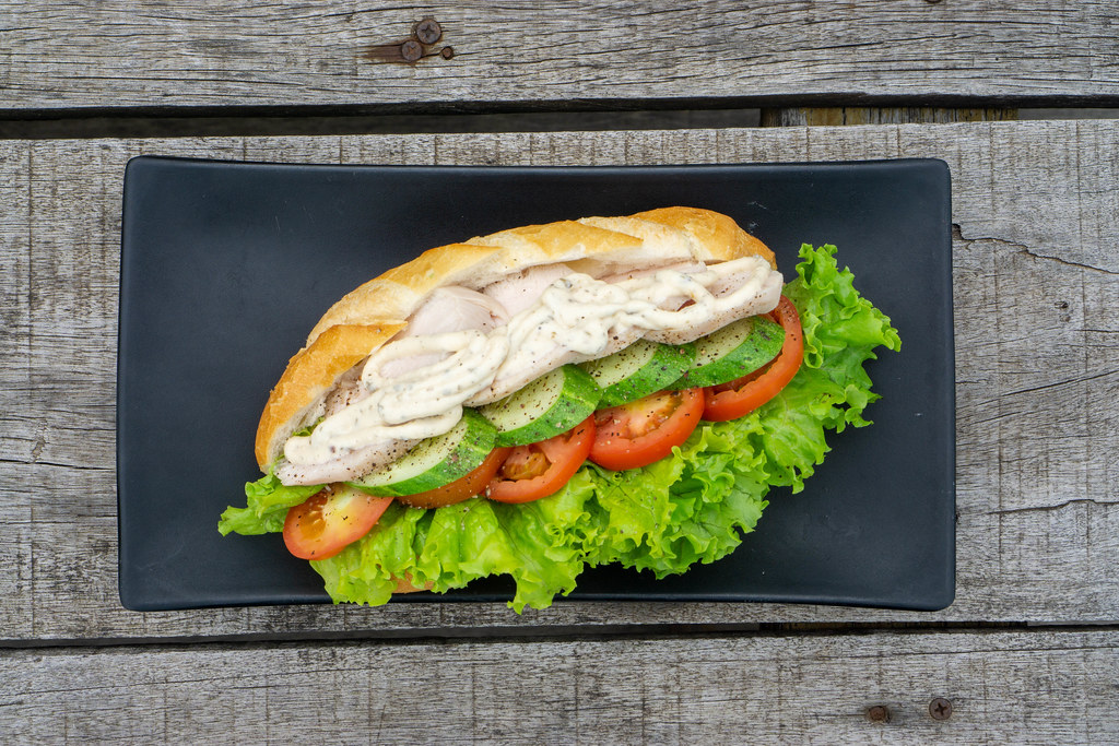 Top View Food Photo of Sandwich with Grilled Chicken Breast, Sliced Cucumber, Tomatoes, Lettuce and Mayonnaise on a Black Plate on a Wooden Table