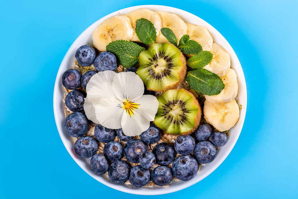 Top view, healthy breakfast, oatmeal with blueberry, banana, mint, kiwi and white flower on blue background