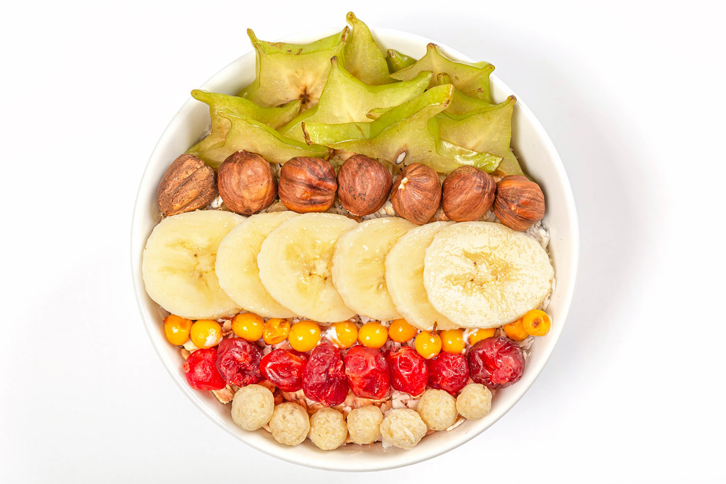 Top view, oatmeal breakfast with fresh fruits, hazelnuts and sun dried cherries on white background