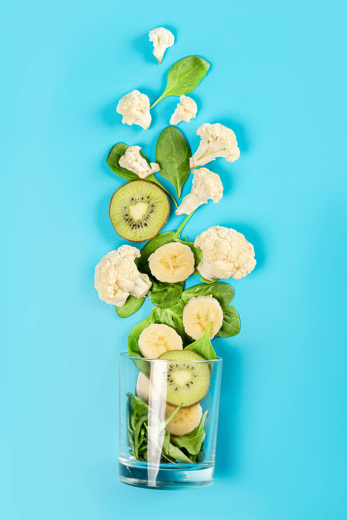 Top view of cauliflower, spinach leaves, banana slices, kiwi and glass on blue background