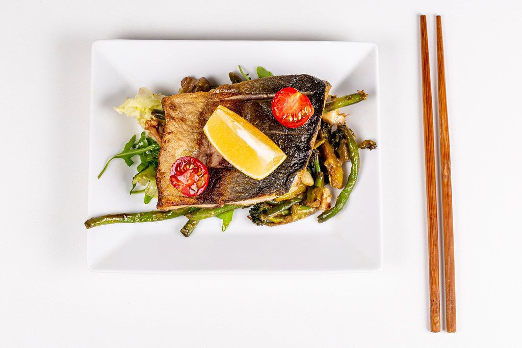 Top view of fried fish with vegetables on white plate with wooden chopsticks