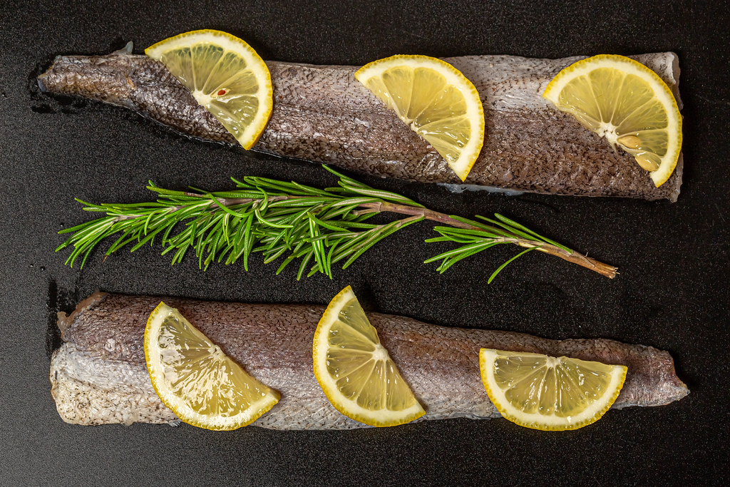 Top view of hake fillet with rosemary sprig and lemon slices on dark background