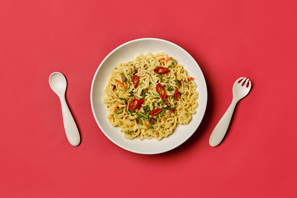 Top view of instant noodle in white bowl with plastic fork and spoon on red background