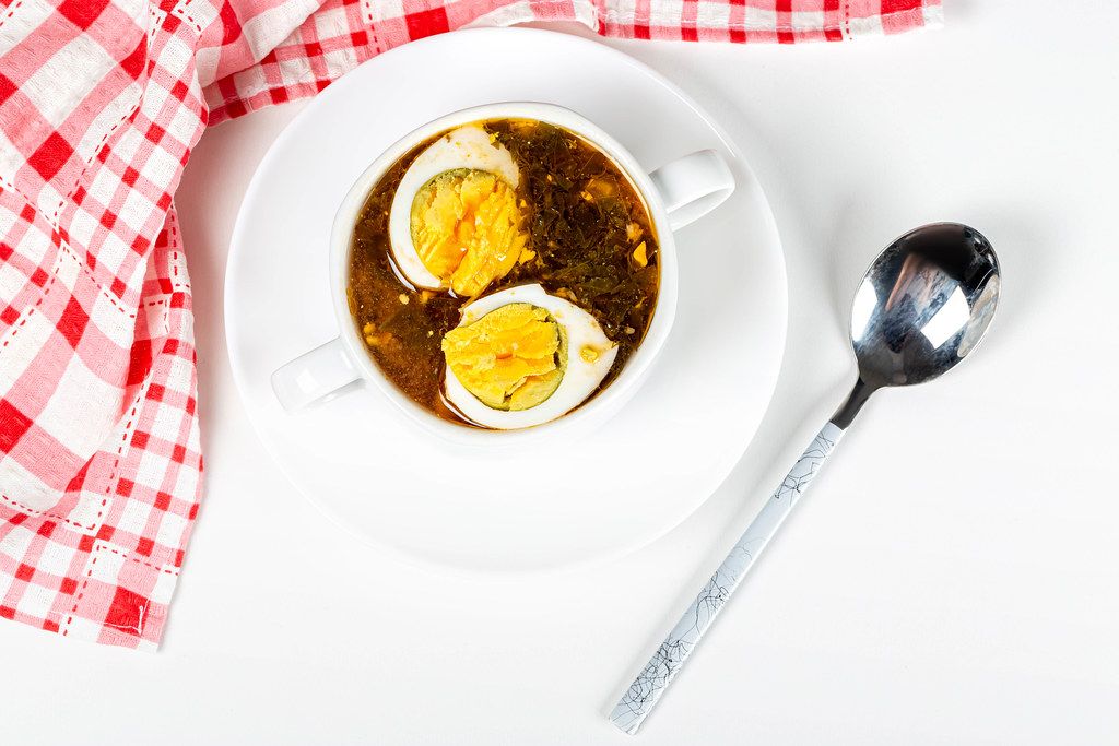 Top view, red soup with herbs and eggs in a tureen on a white plate with a spoon and a kitchen towel