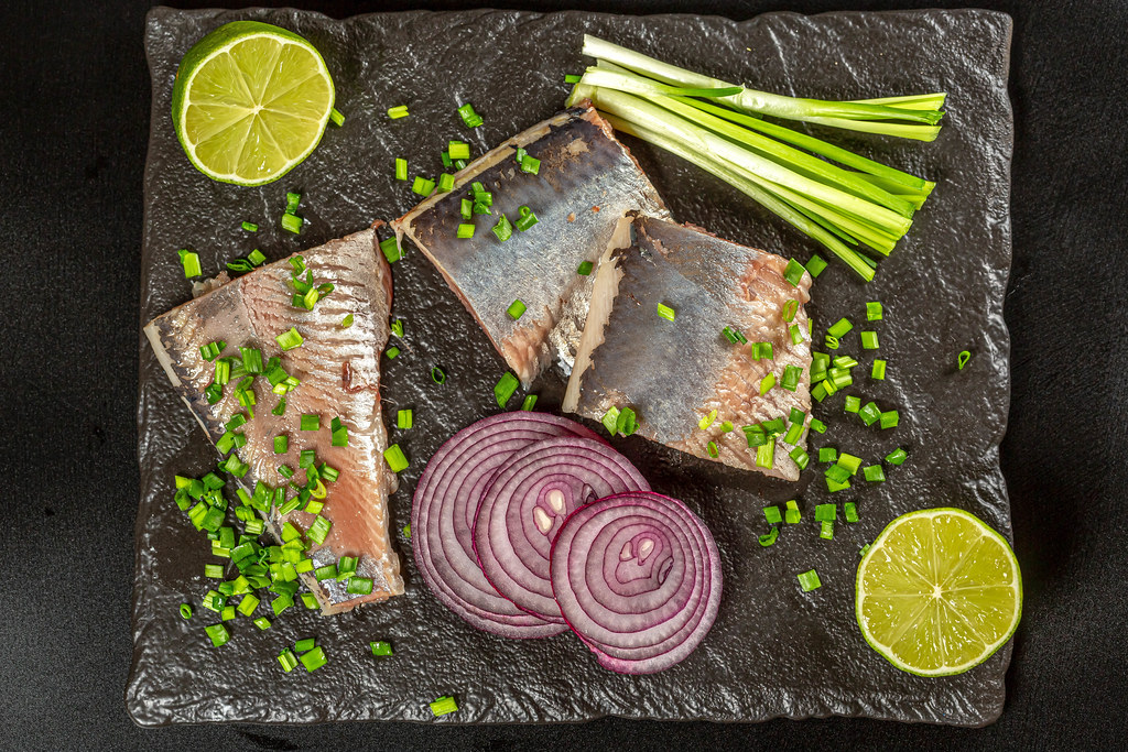 Top view, slices of marinated herring fillet with lime, green and purple onions on black stone tray