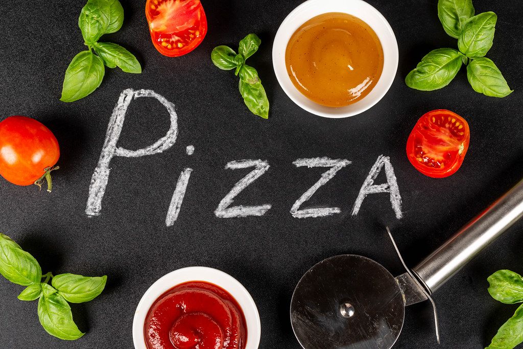 Top view, the inscription pizza on a black background with sauces, basil, tomatoes and a knife