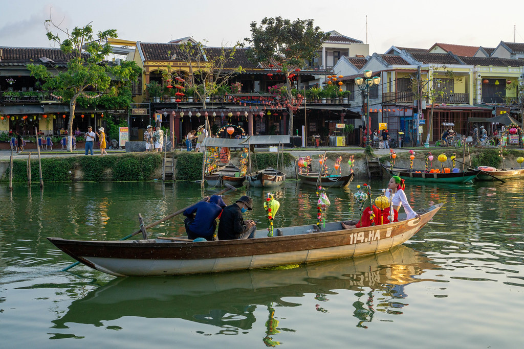 Touristen auf einem Ruderboot mit bunten Laternen auf dem Thu Bon Fluss mit Restaurants und Shops im Hintergrund in der Altstadt von Hoi An, Vietnam